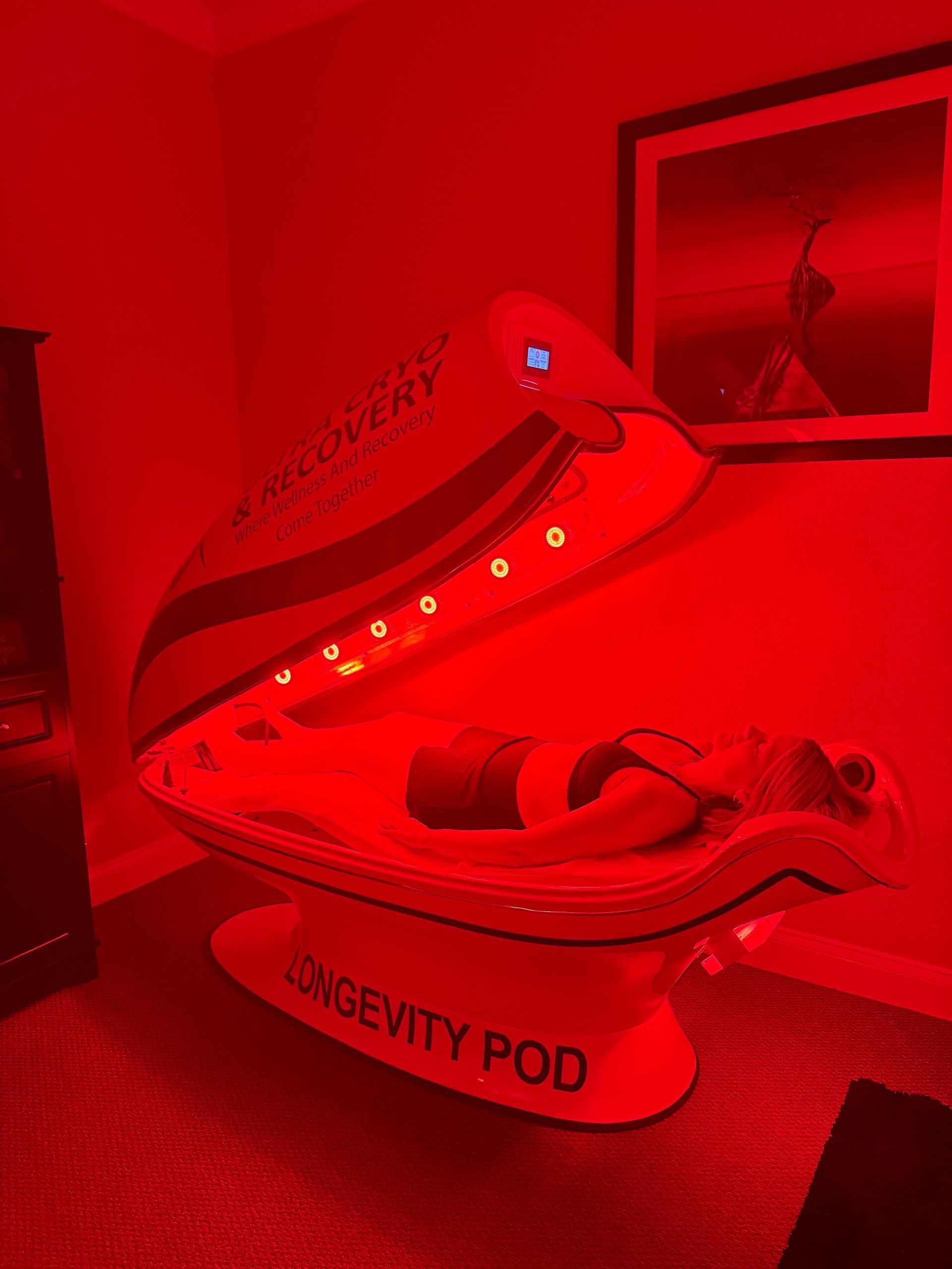 A person reclines inside a Longevity Pod emitting bright red light in a dimly lit, red-toned room.