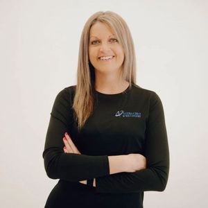 Woman with arms crossed, smiling, wearing a black shirt with a small logo, set against a white background.