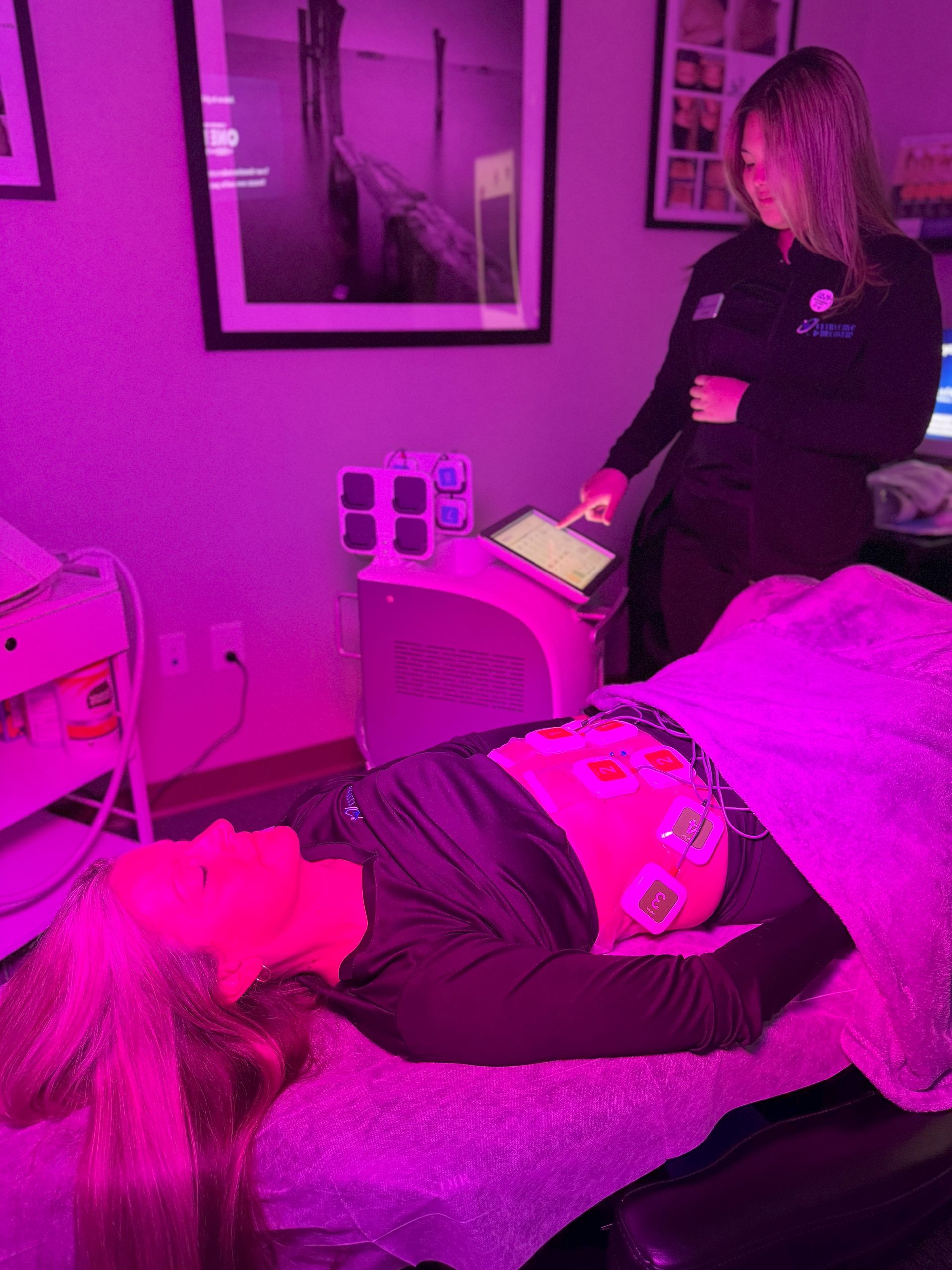 Woman receiving facial treatment under pink light; technician operates tablet in spa.