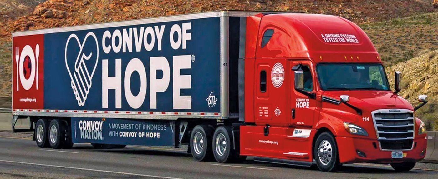 Red semi-truck hauling a trailer with "Convoy of Hope" text and heart logo on highway.