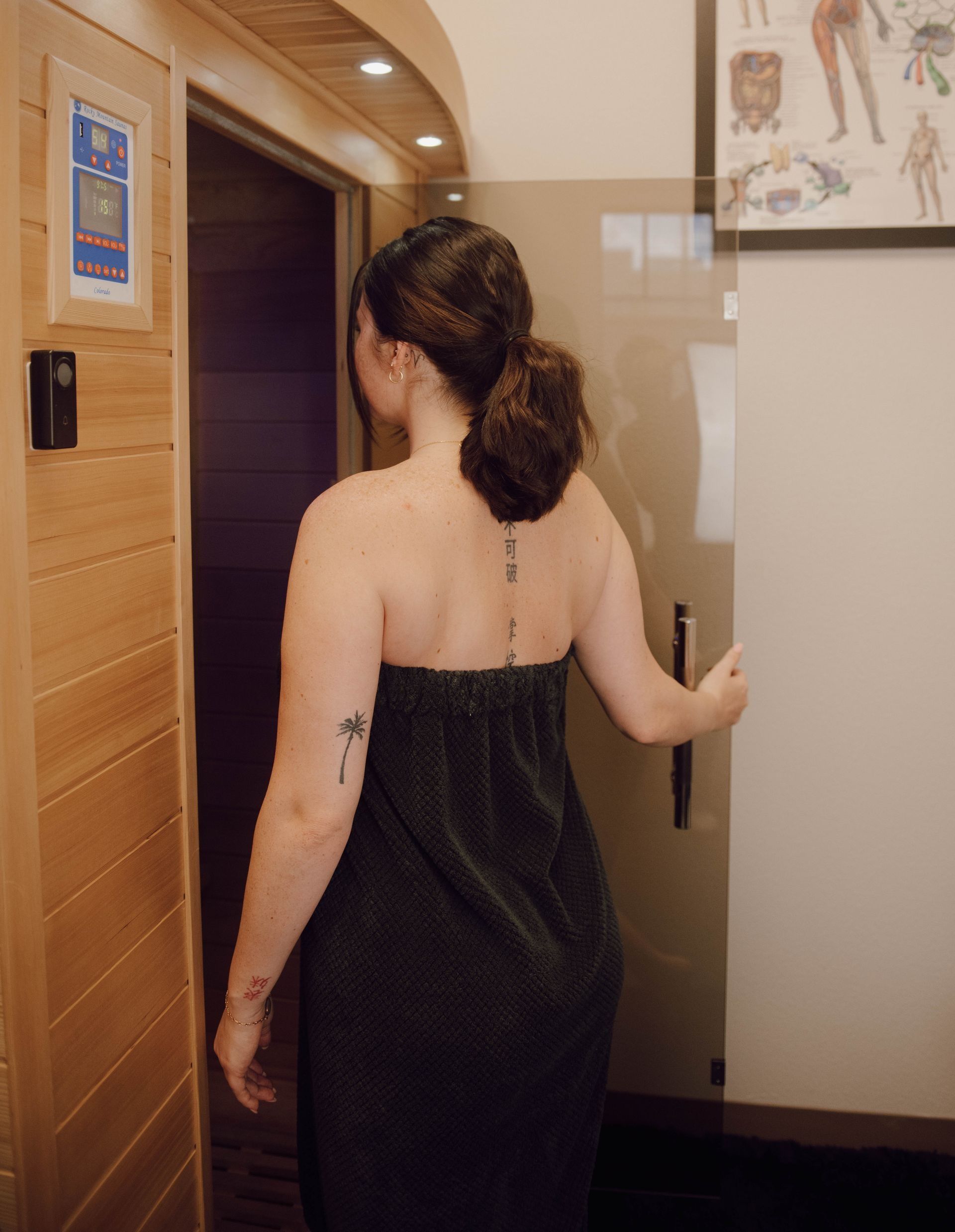 Person in a towel entering a sauna, opening a glass door, with wood paneling and controls.