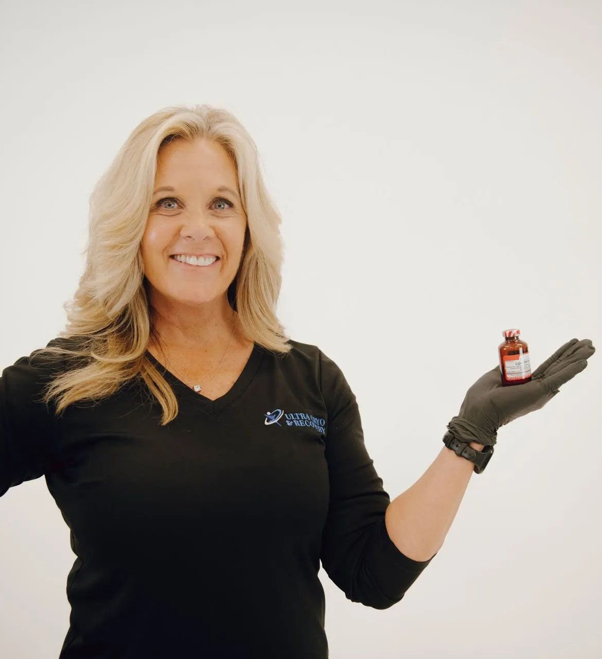 Woman in black shirt and gloves holding small bottle, smiling. Bright background.