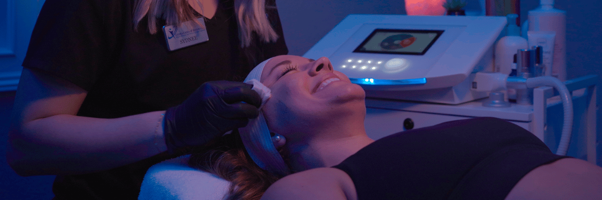 Person receiving facial treatment at a clinic. Technician wearing gloves applies product. Blue lighting.