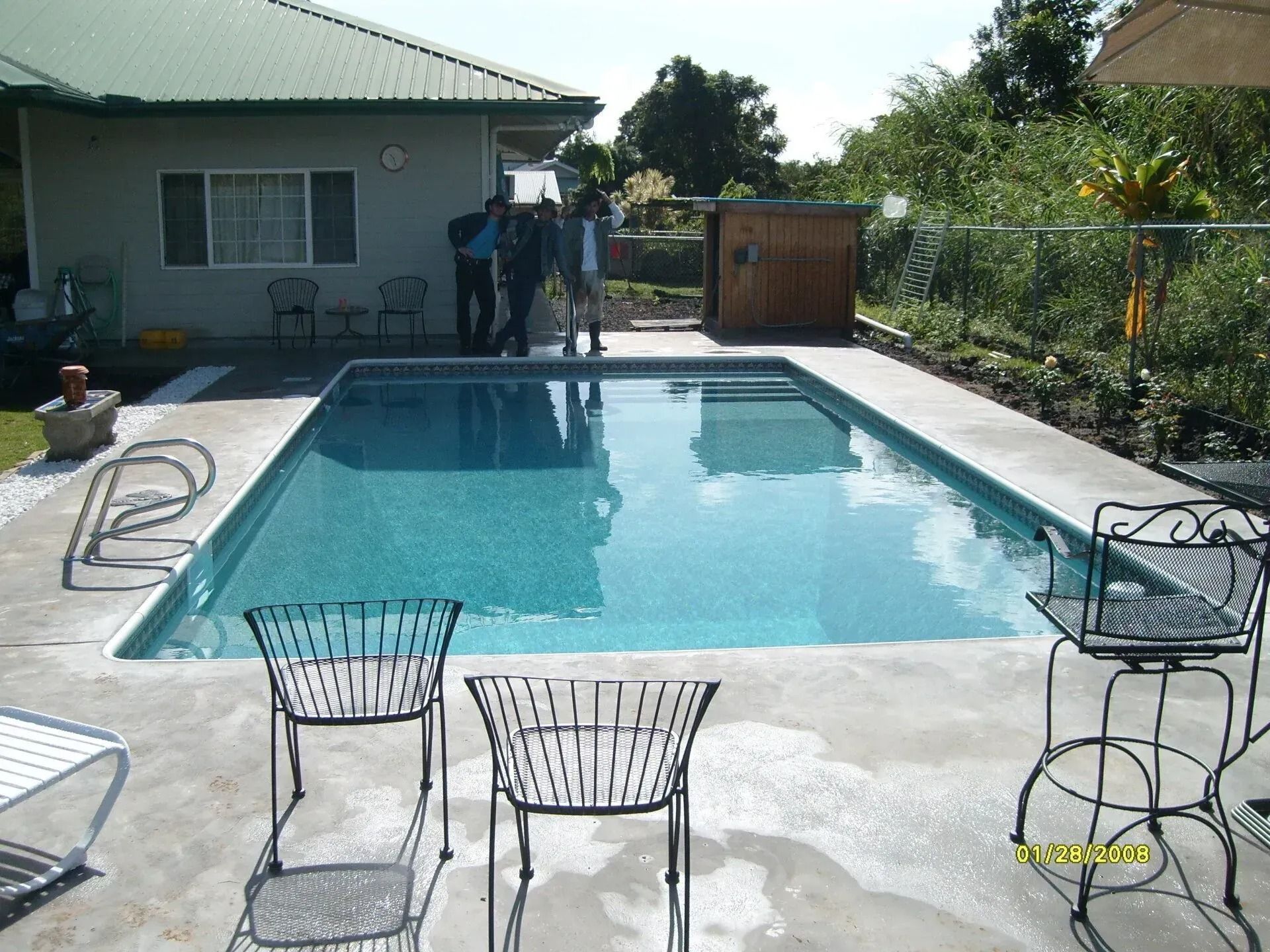 Backyard swimming pool with patio chairs, a house, and a wooden fence on a sunny day