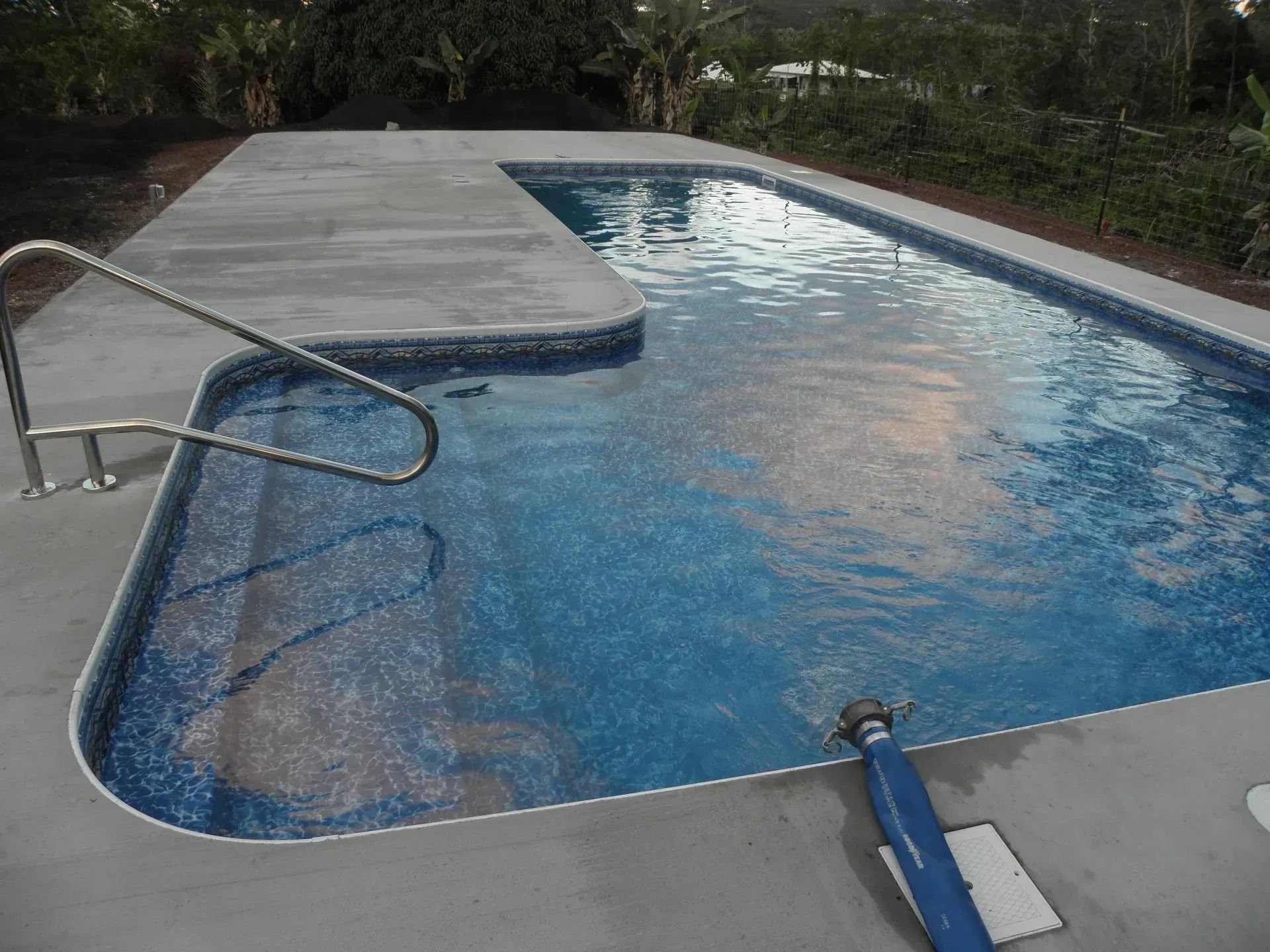 Outdoor swimming pool with a diving board, metal ladder, and surrounding concrete deck, bordered by trees.