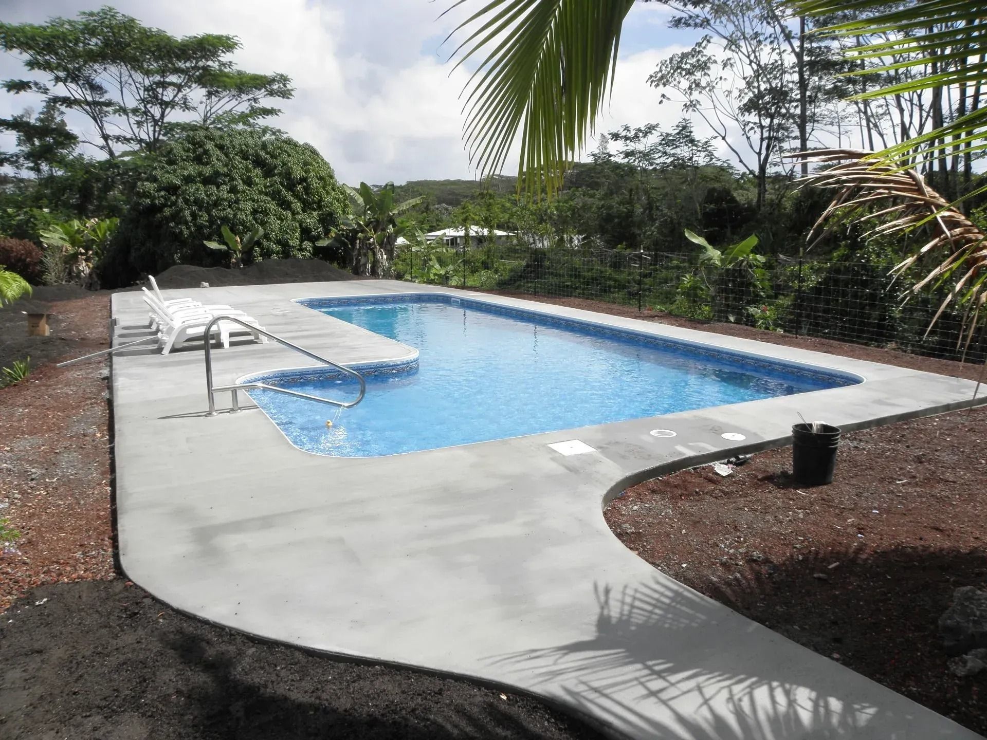 Backyard swimming pool with white deck, surrounded by tropical greenery and cloudy sky.