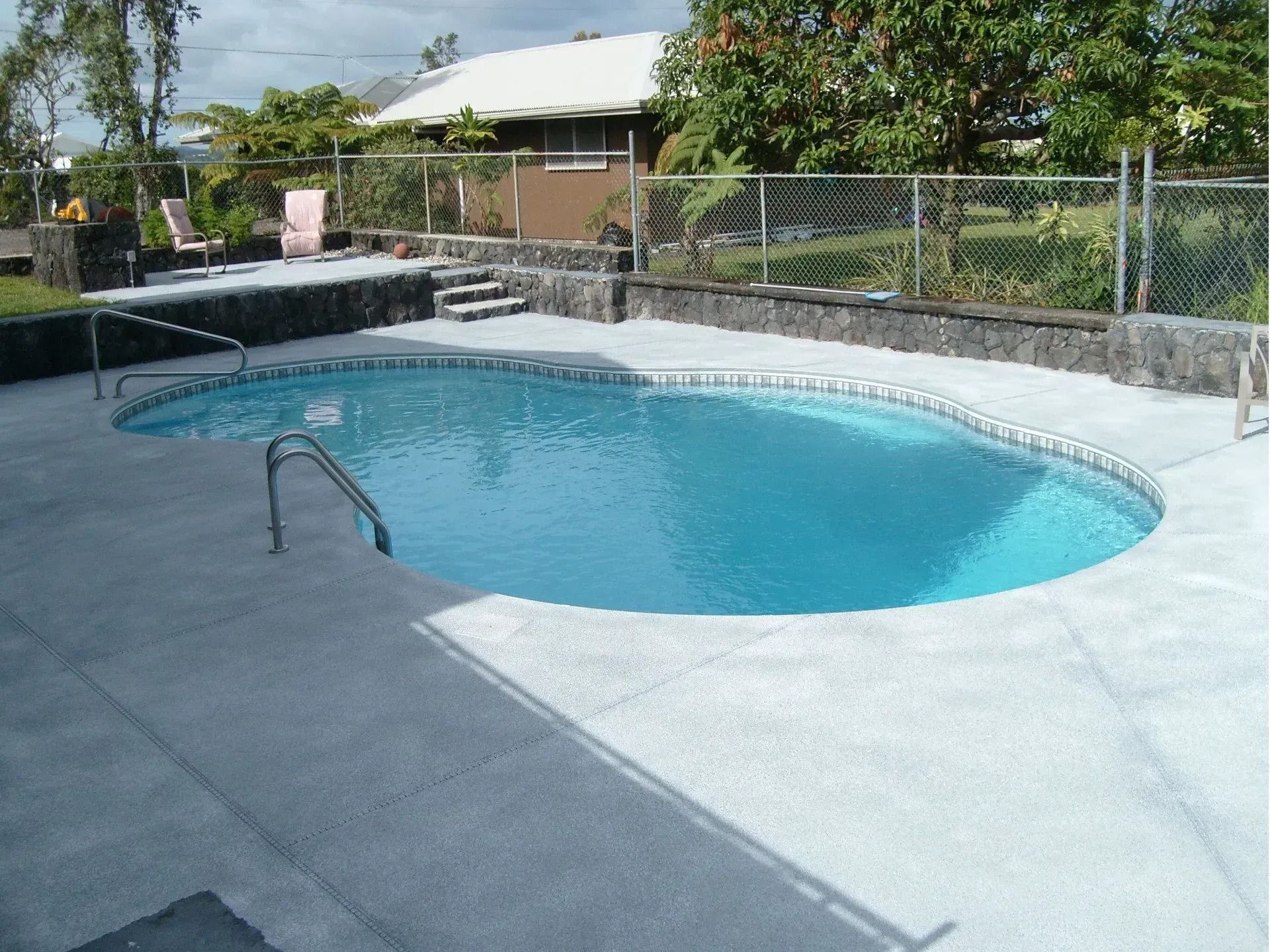 Backyard swimming pool with blue water and a concrete deck, surrounded by a chain-link fence and trees
