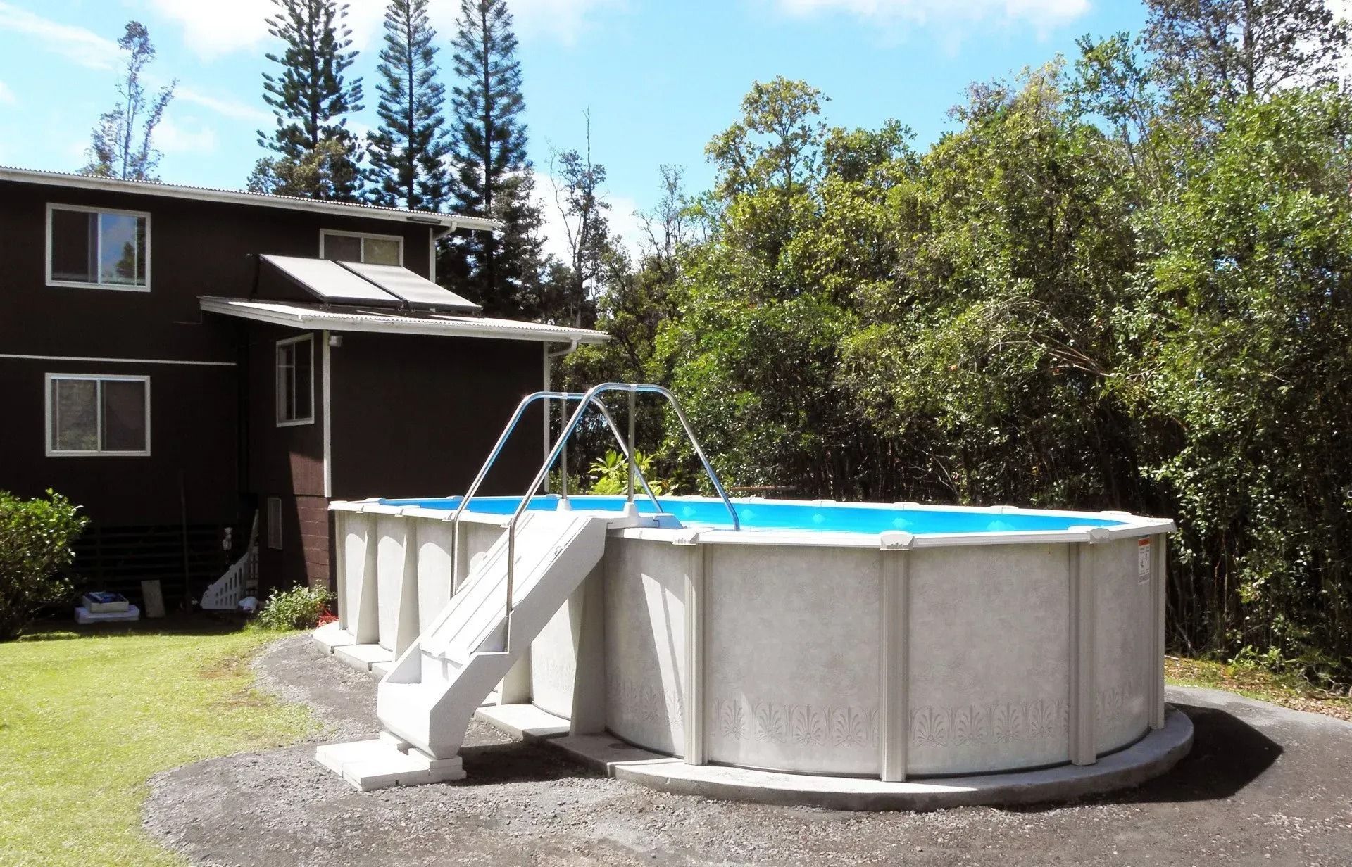 Above-ground pool with a white ladder in a sunny backyard beside a house and trees