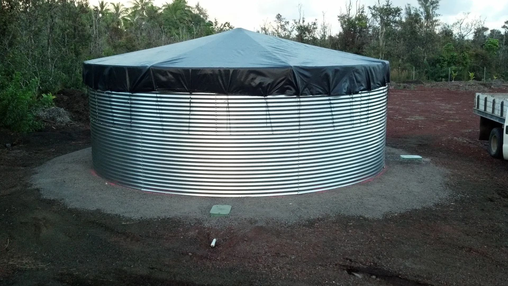Round corrugated metal water tank with black cover on gravel in an outdoor yard