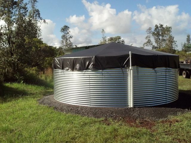 Round metal water tank with black cover in a grassy field under a cloudy sky