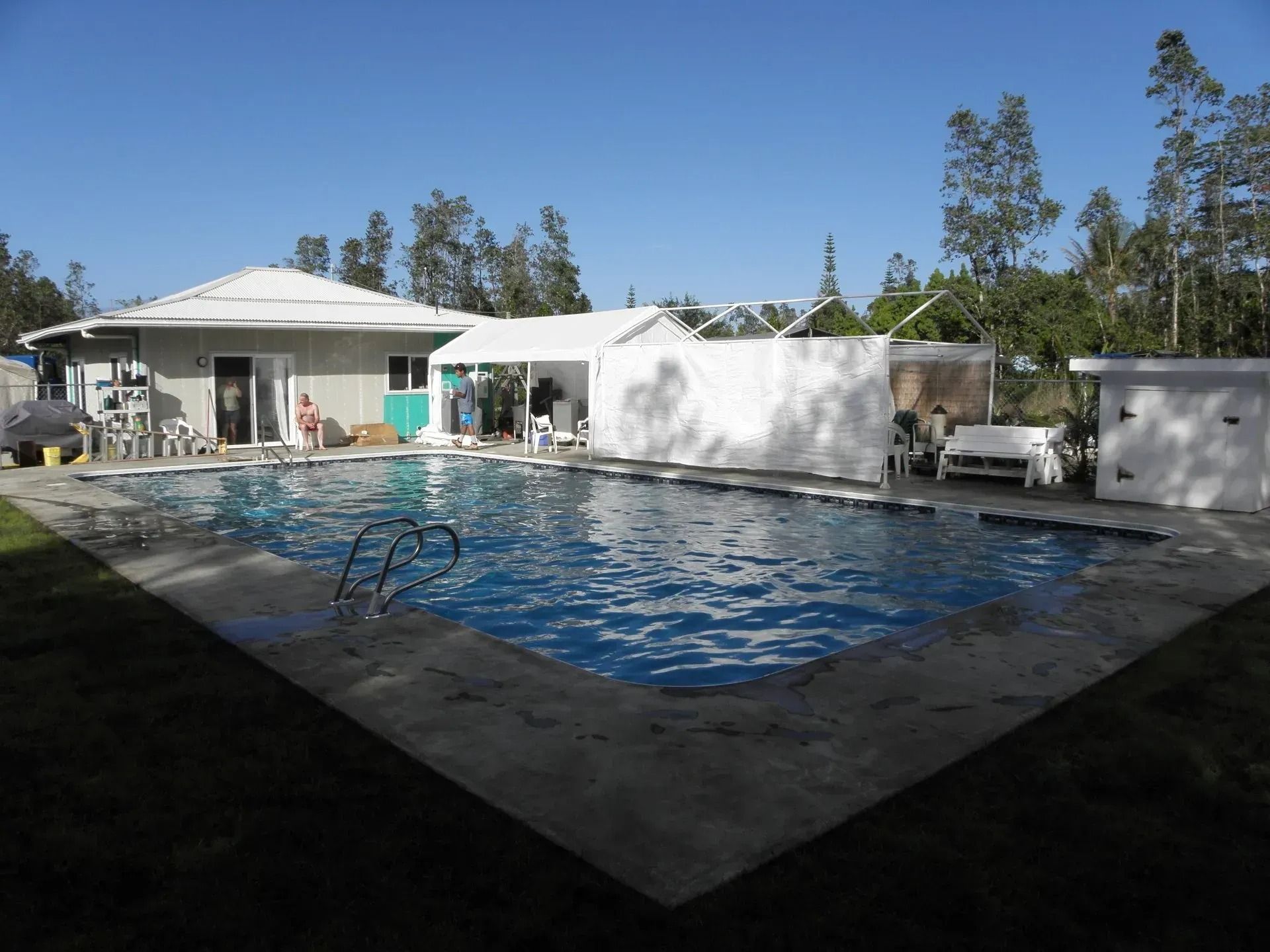 Backyard pool beside a white house on a sunny day, with trees and lounge chairs nearby