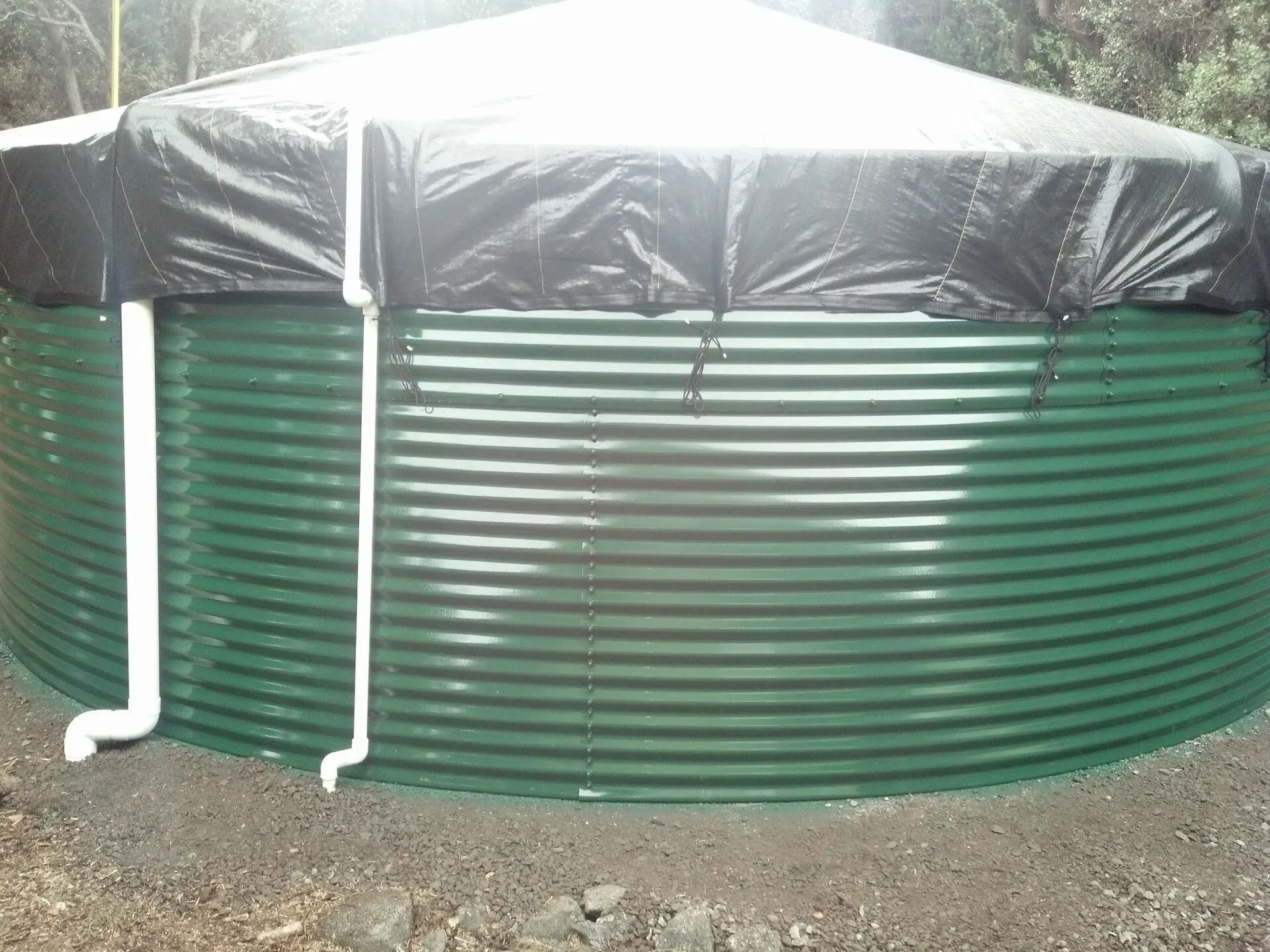 Green corrugated water tank with black tarp cover and white downspouts outdoors