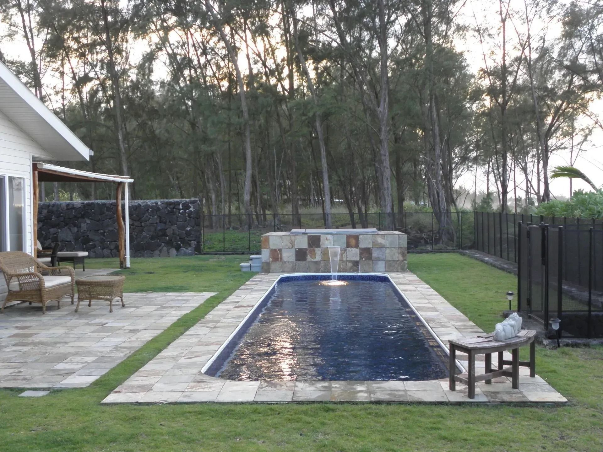 Backyard pool with stone deck, patio chairs, and trees at dusk