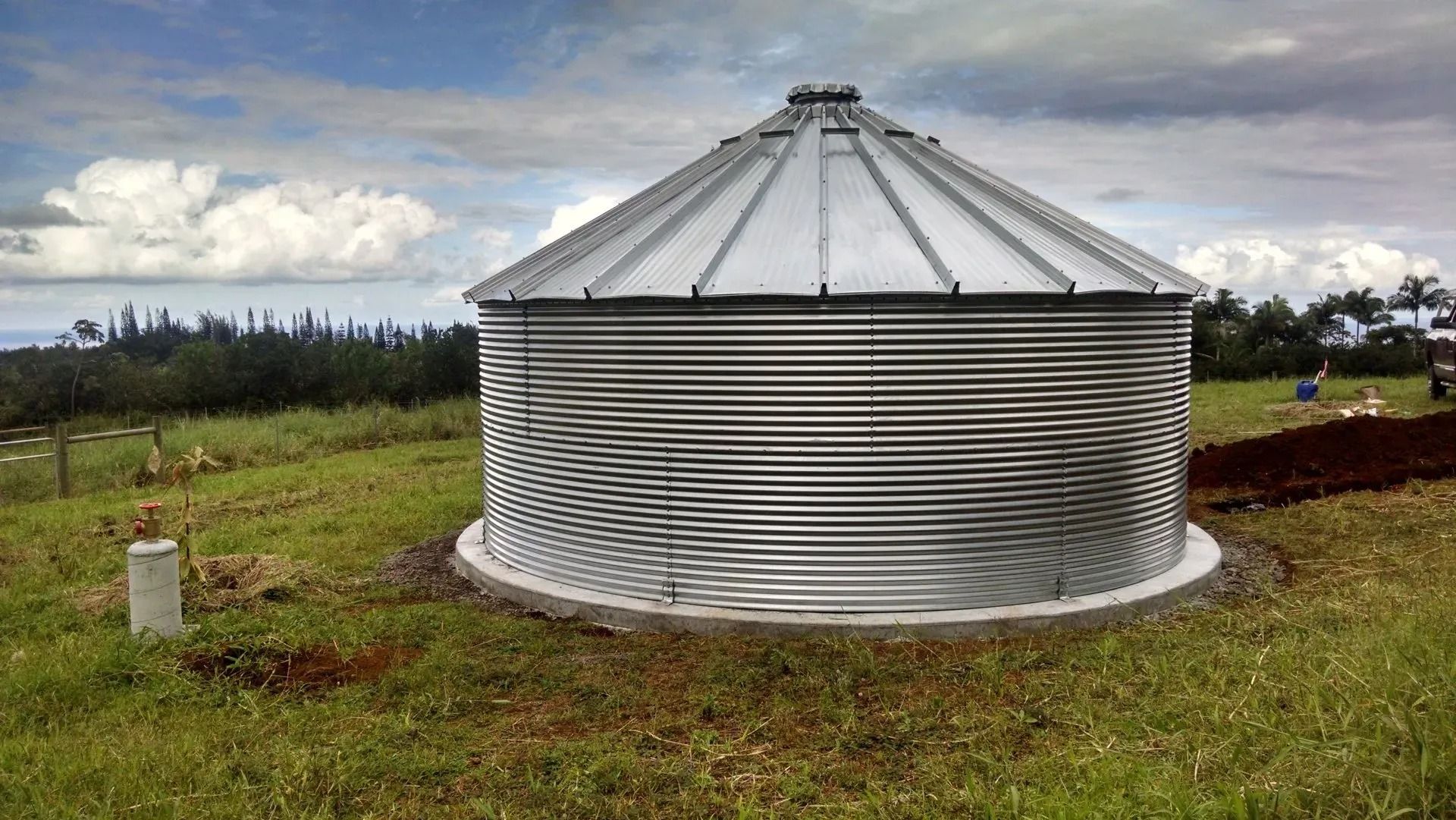 Metal grain silo in a grassy field under a cloudy sky, with a farm fence in the background