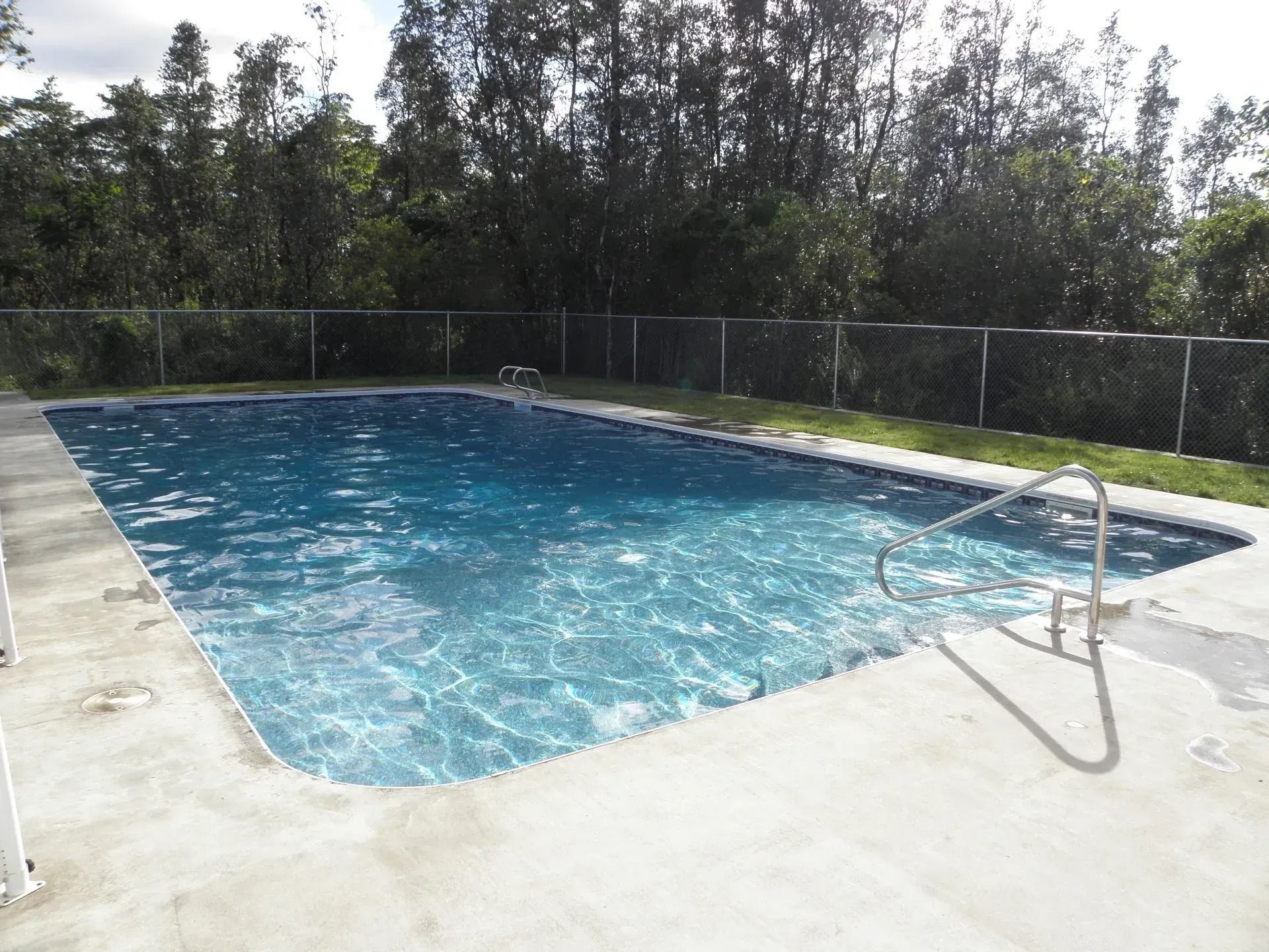 Outdoor swimming pool with blue water, surrounded by a concrete deck and fenced by trees.