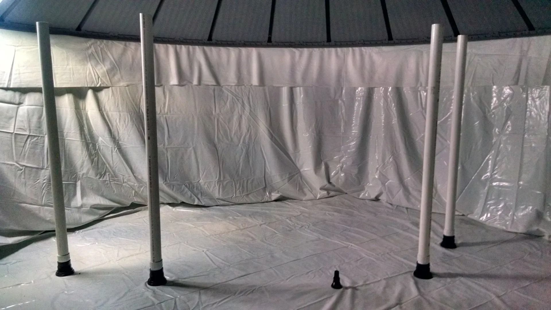White tarp-covered room with several vertical poles and a central hanging cord or weight on the floor.