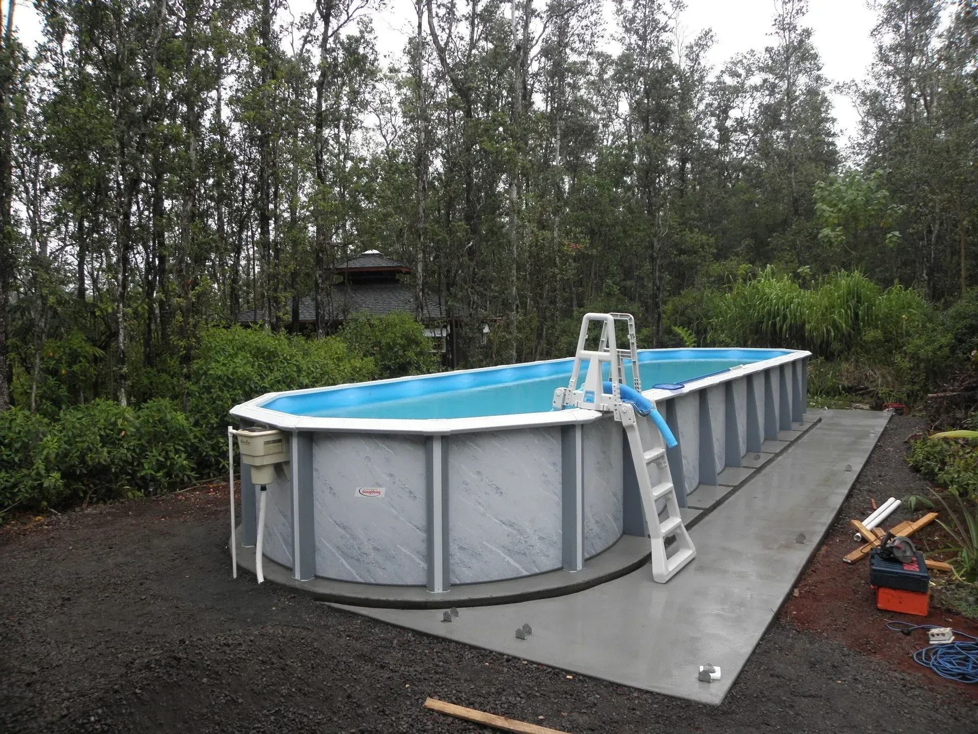 Above-ground pool on a gravel pad with ladder, surrounded by trees and bushes