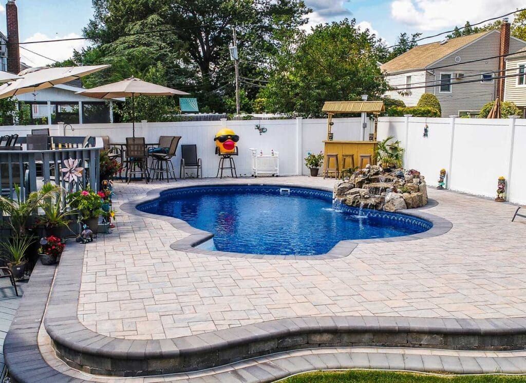 Backyard patio with a blue swimming pool, pavers, umbrella table, and white privacy fence