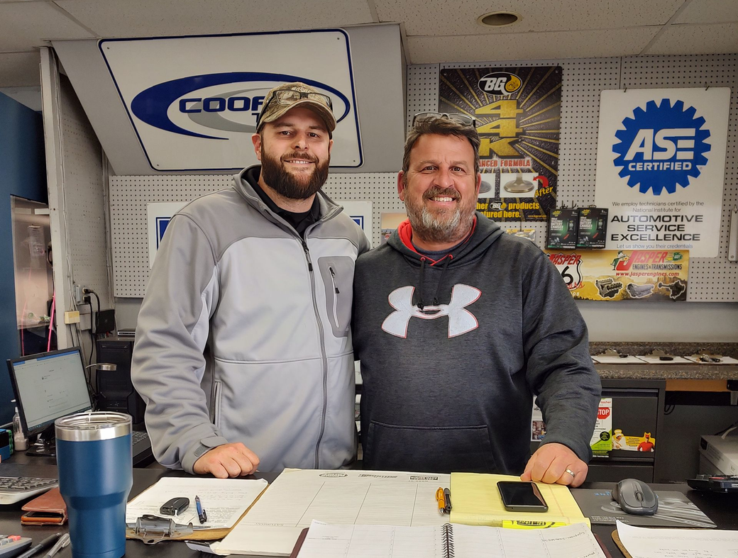 Two men standing behind a counter in an auto shop, smiling with papers and tools on the counter.