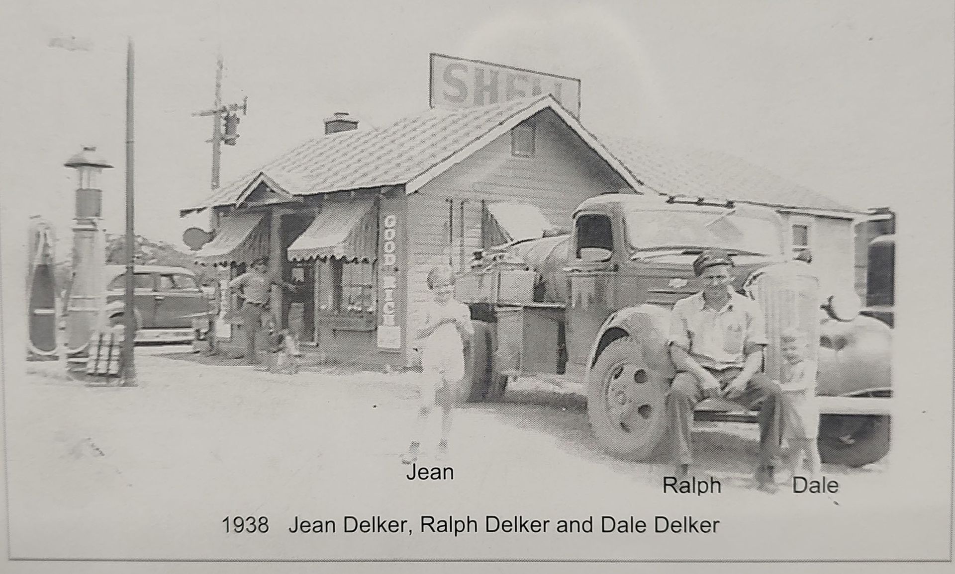 Vintage black-and-white photo of a tractor parked beside a small rustic building with people standing nearby.