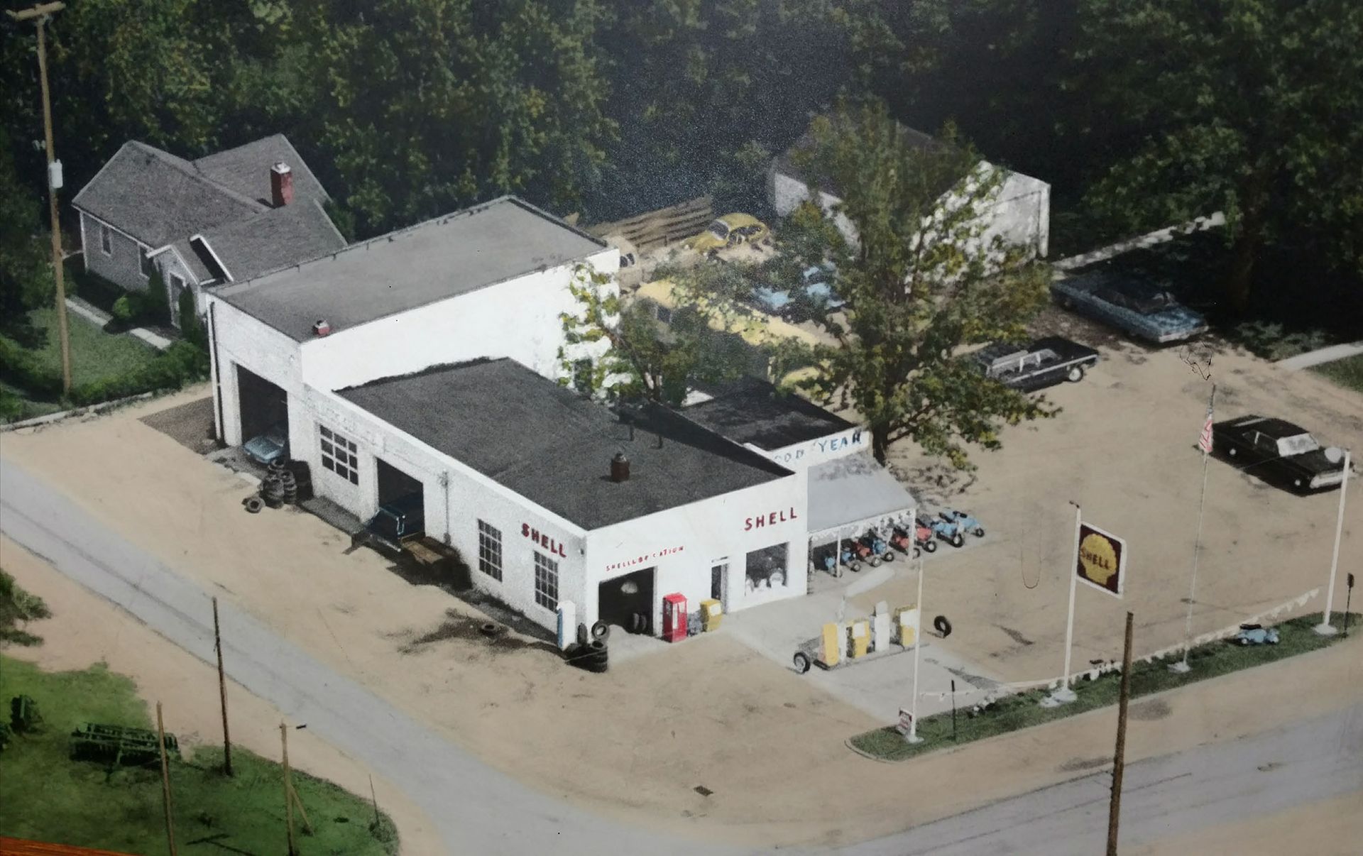 Aerial view of a white building with a dark roof beside a dirt lot and a tree-lined road.