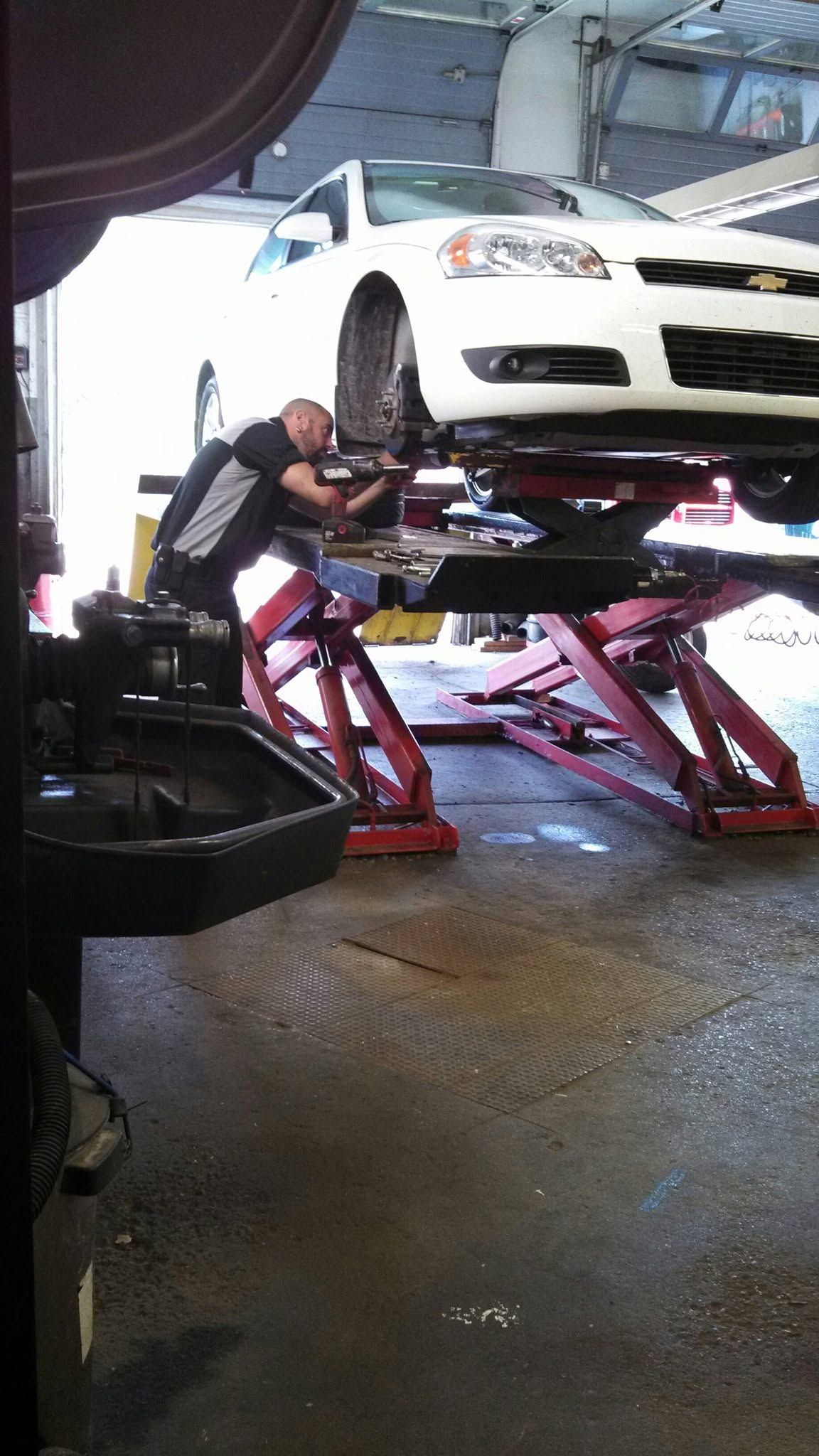 Mechanic working under a white car lifted on a red hydraulic lift in an auto shop