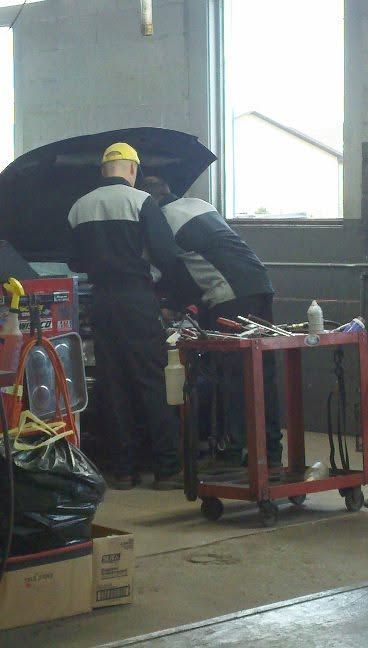 Two workers in a garage inspect a car under an open hood near tool carts and equipment.
