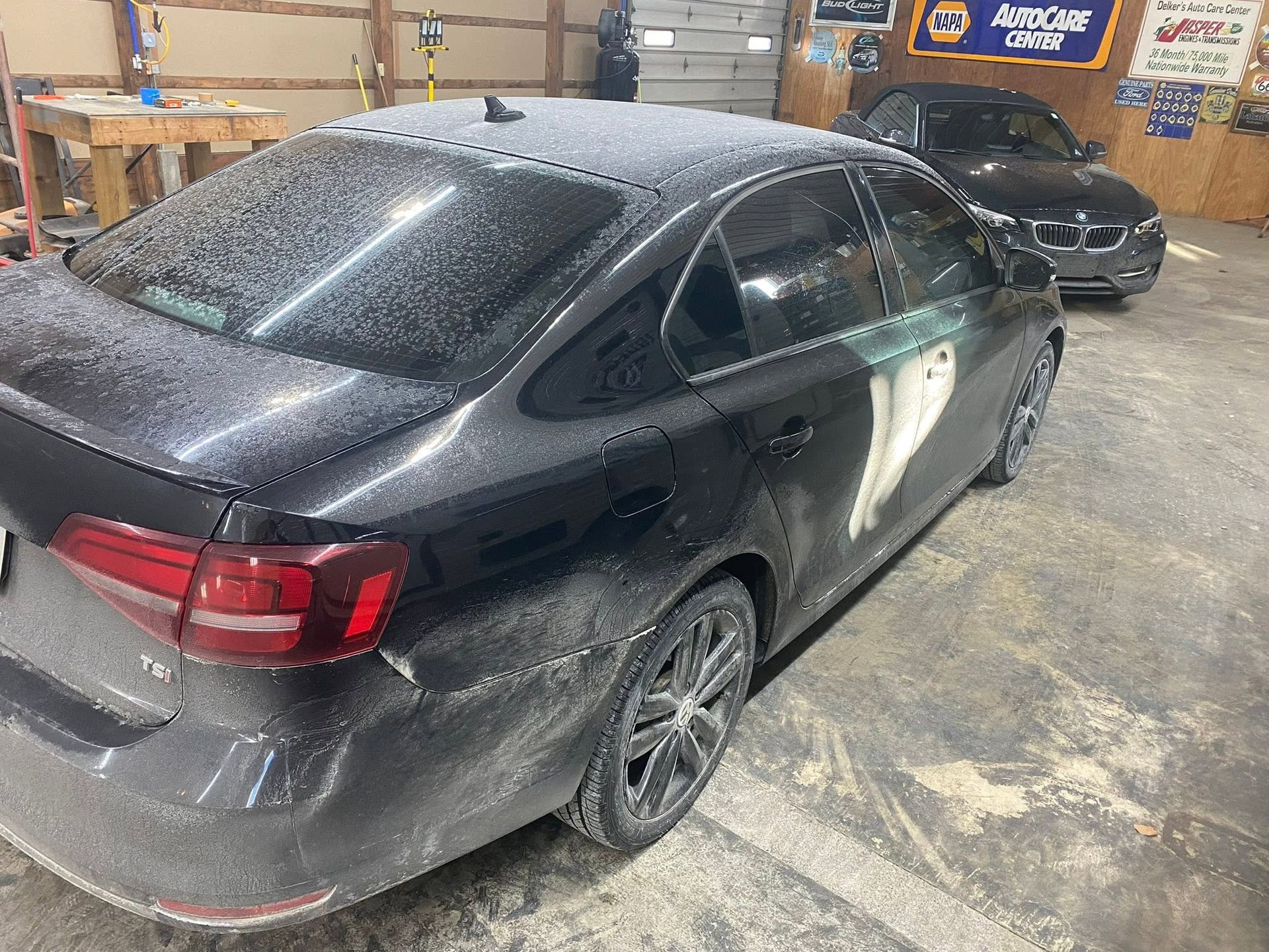Two dark sedans parked in a dusty garage, with one covered in dust and damage on the side.