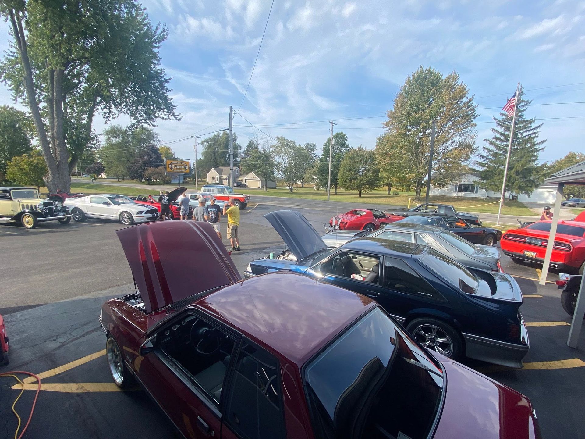 Car meet in a parking lot with several sports cars, some with hoods open, under a sunny sky.