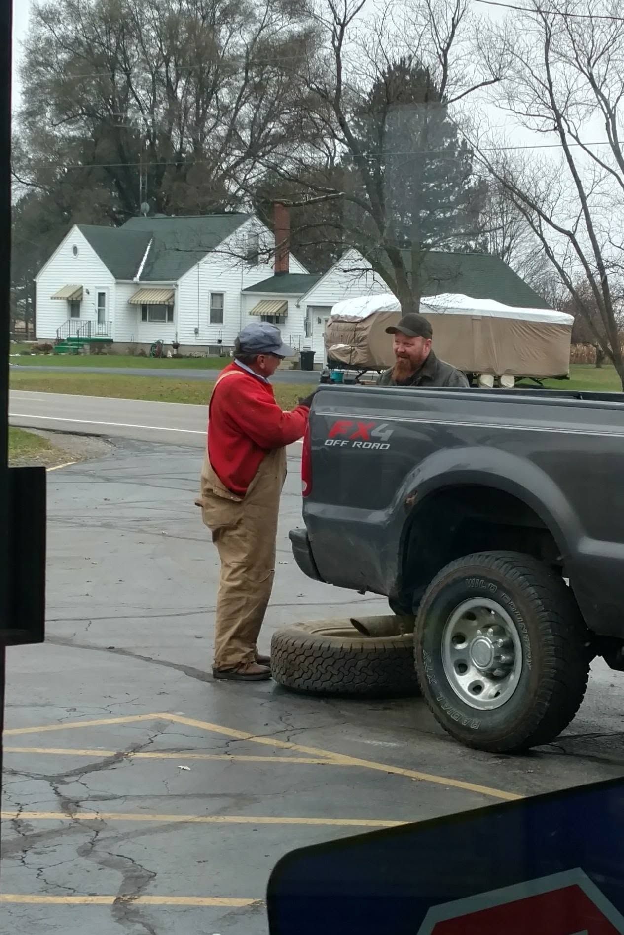 Two men talking beside a gray pickup truck with a spare tire on a wet roadside in a residential area