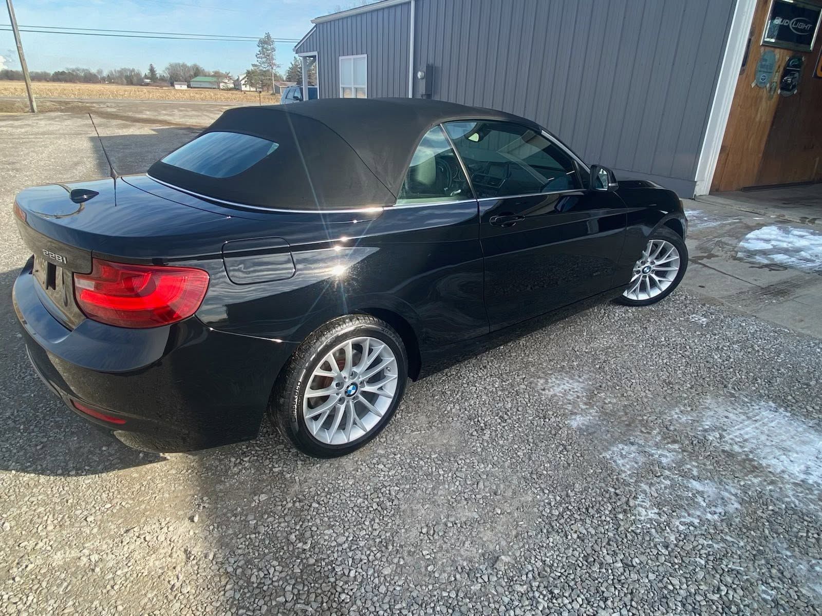 Black BMW convertible parked on a gravel lot beside a building, with snow patches nearby.
