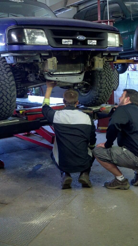 Mechanics inspecting the front underside of a lifted truck in a garage, with one using a light.
