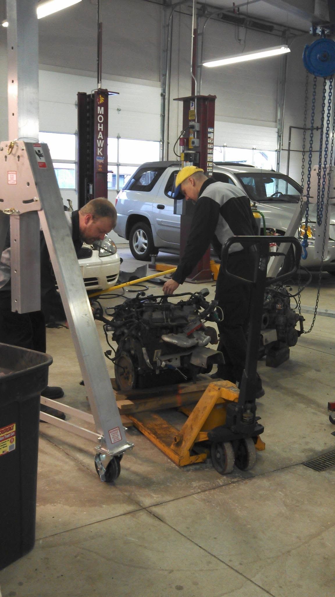Two mechanics working on a car engine in an auto repair shop with a lifted vehicle in the background