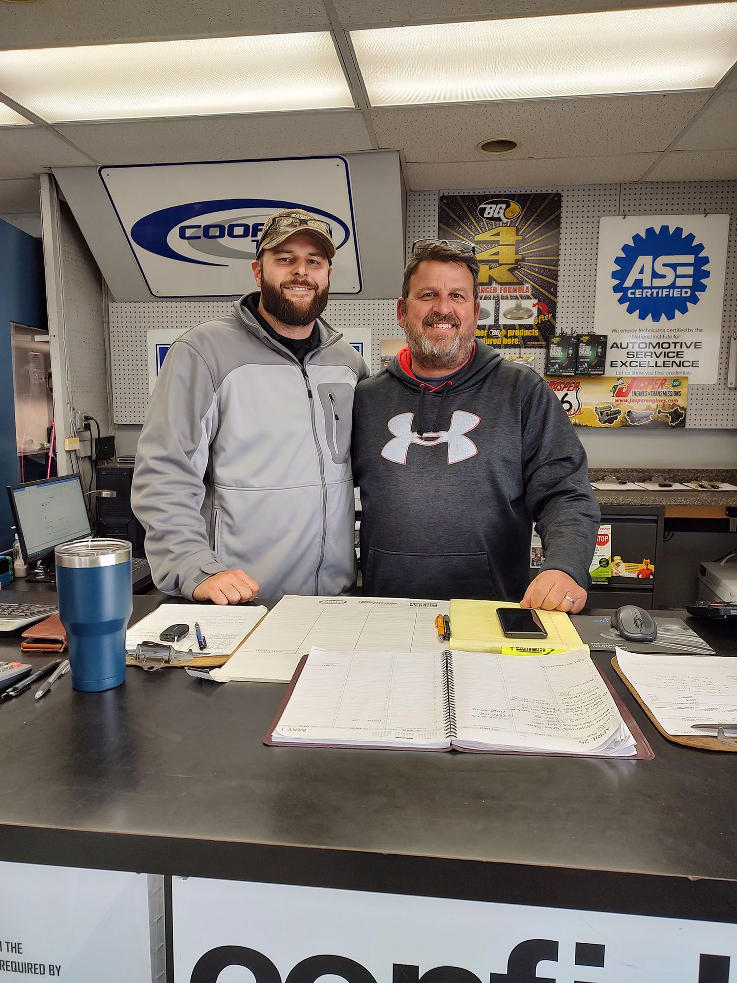 Two men standing behind a counter in an auto shop, with papers, a tumbler, and shop signs in the background.