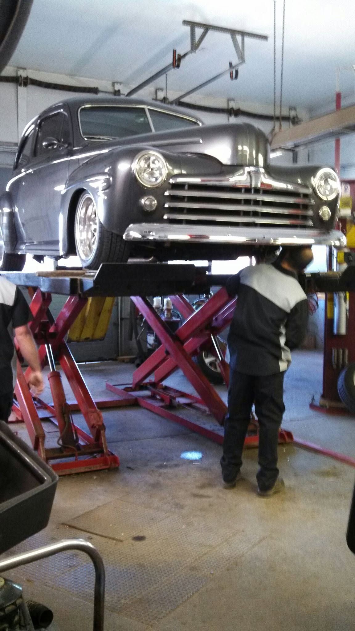 Vintage car raised on a hydraulic lift in a garage, with a mechanic working underneath.