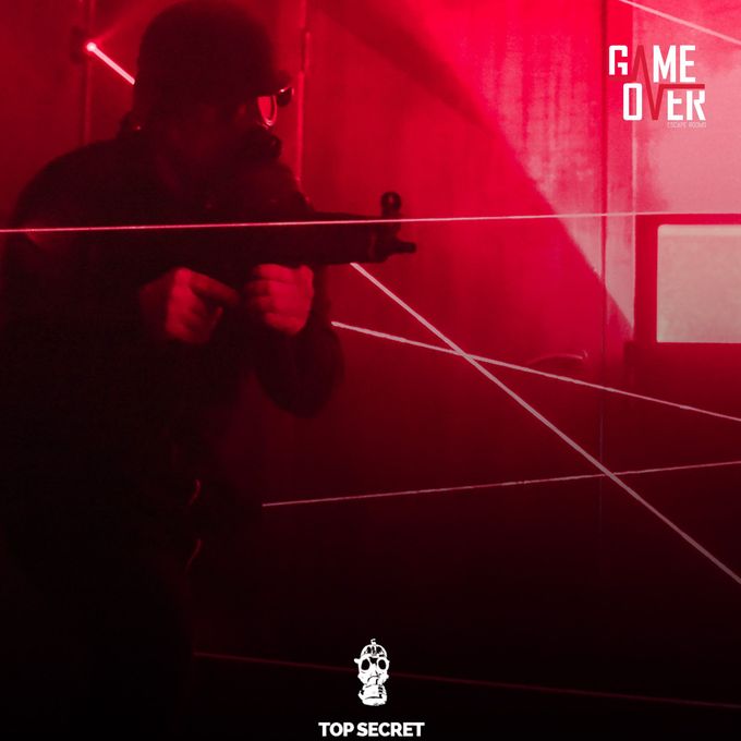 A person dressed in dark tactical gear, including a helmet and goggles, is holding a firearm while navigating through a room filled with red laser beams. The room is dimly lit with a red hue, and the lasers crisscross the space. At the bottom of the image, there is an icon of a skull wearing headphones above the text 'TOP SECRET.