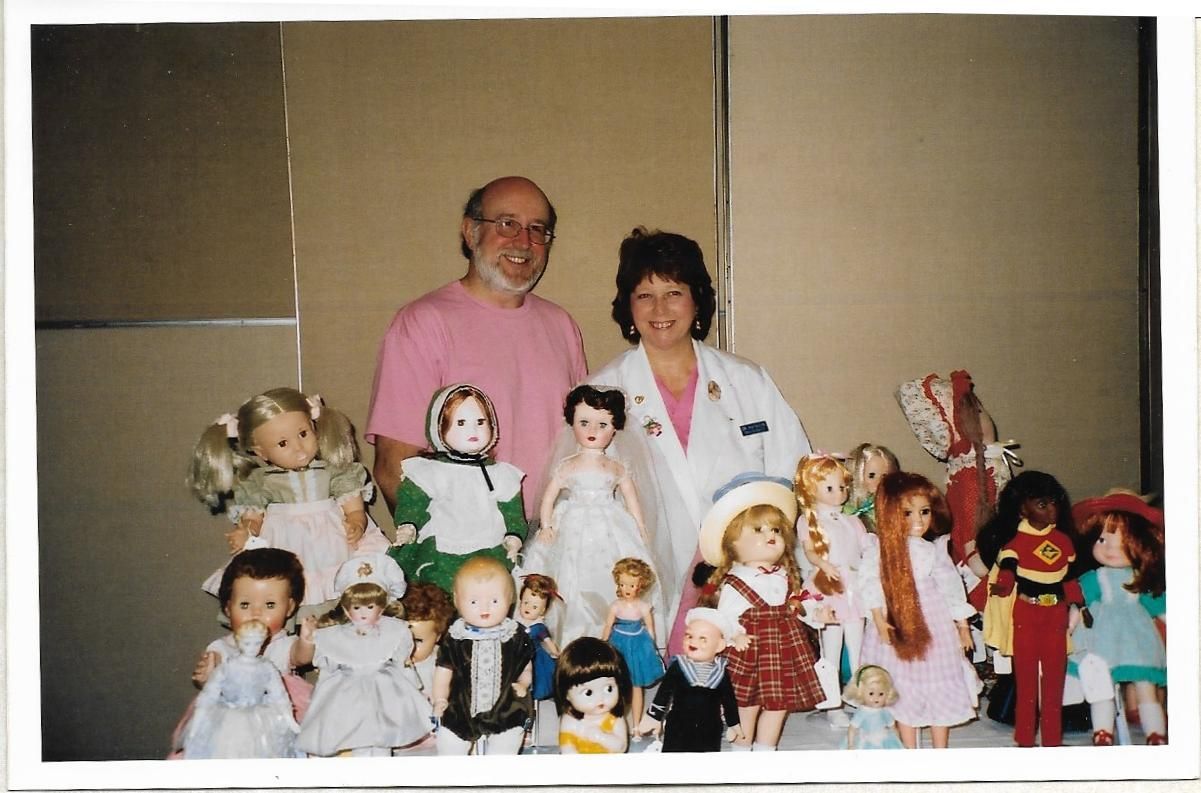 Woman in lab coat examines doll's arm with tools at a white table. A bucket of brushes and doll parts are nearby.