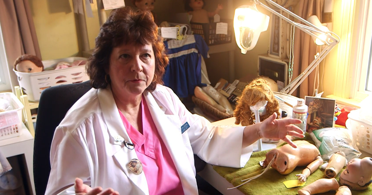 Woman in lab coat examines doll's arm with tools at a table.