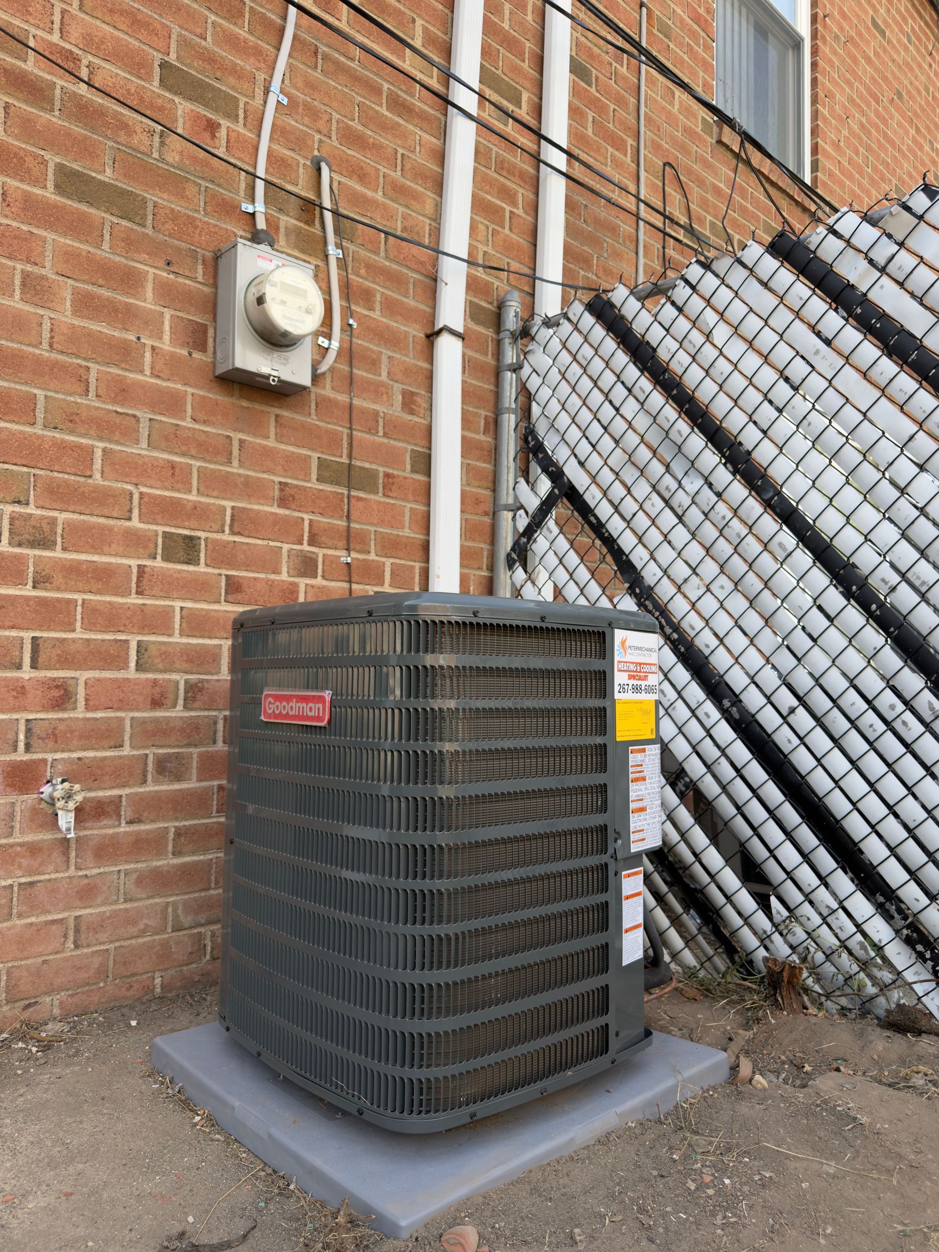 Outdoor HVAC unit beside a brick wall, with a chain-link fence and stacked materials nearby.
