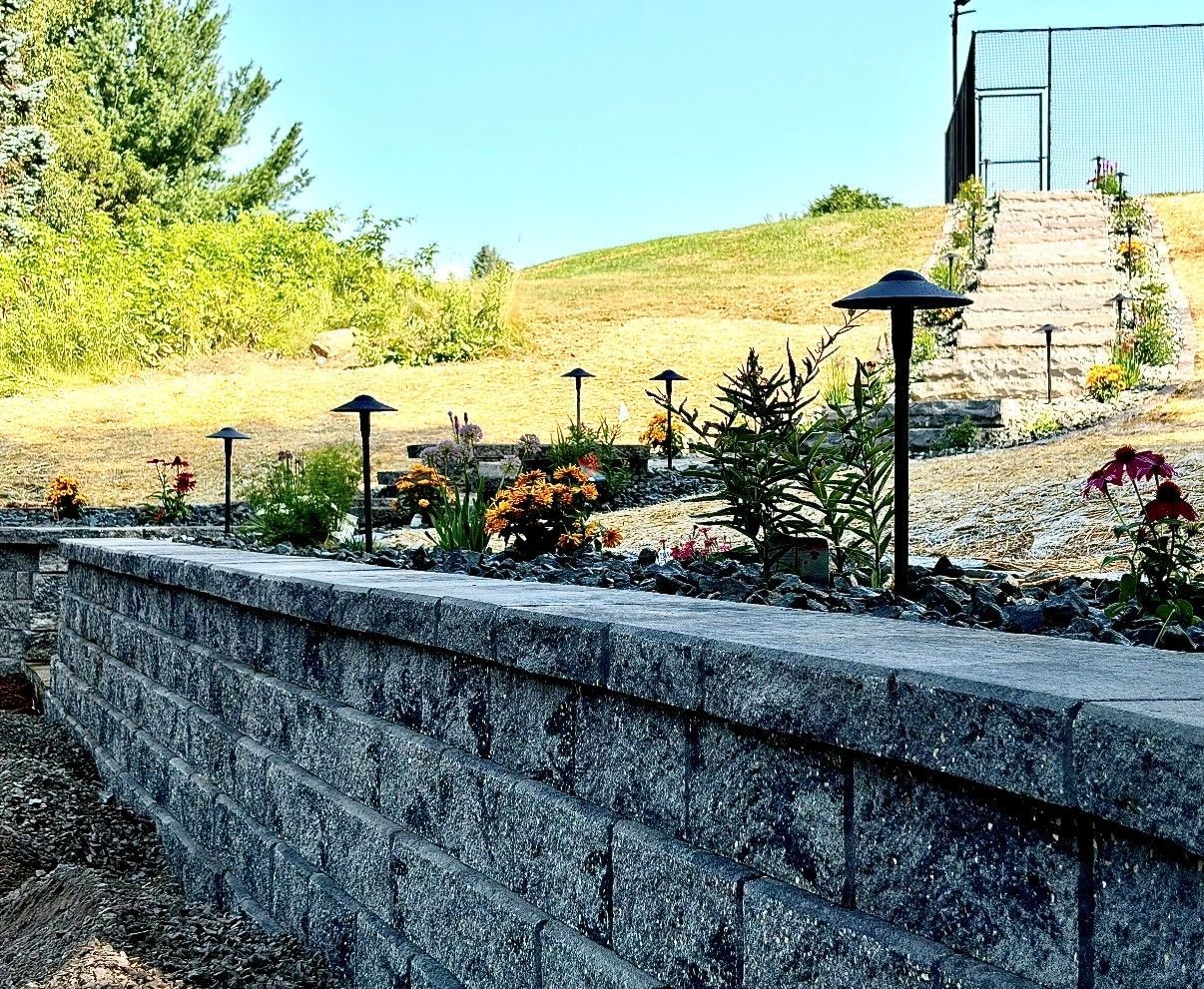 Stone retaining wall with flowerbed and black path lights beside grassy steps under a bright blue sky