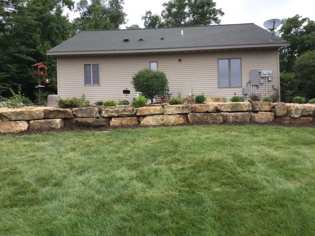 a house with a stone wall in front of it and a lush green lawn .