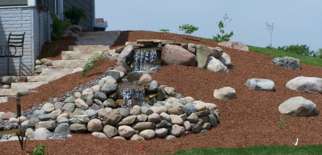 a small waterfall is surrounded by rocks and mulch .