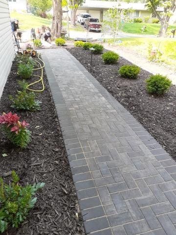 a brick walkway leading to a house surrounded by plants and mulch .