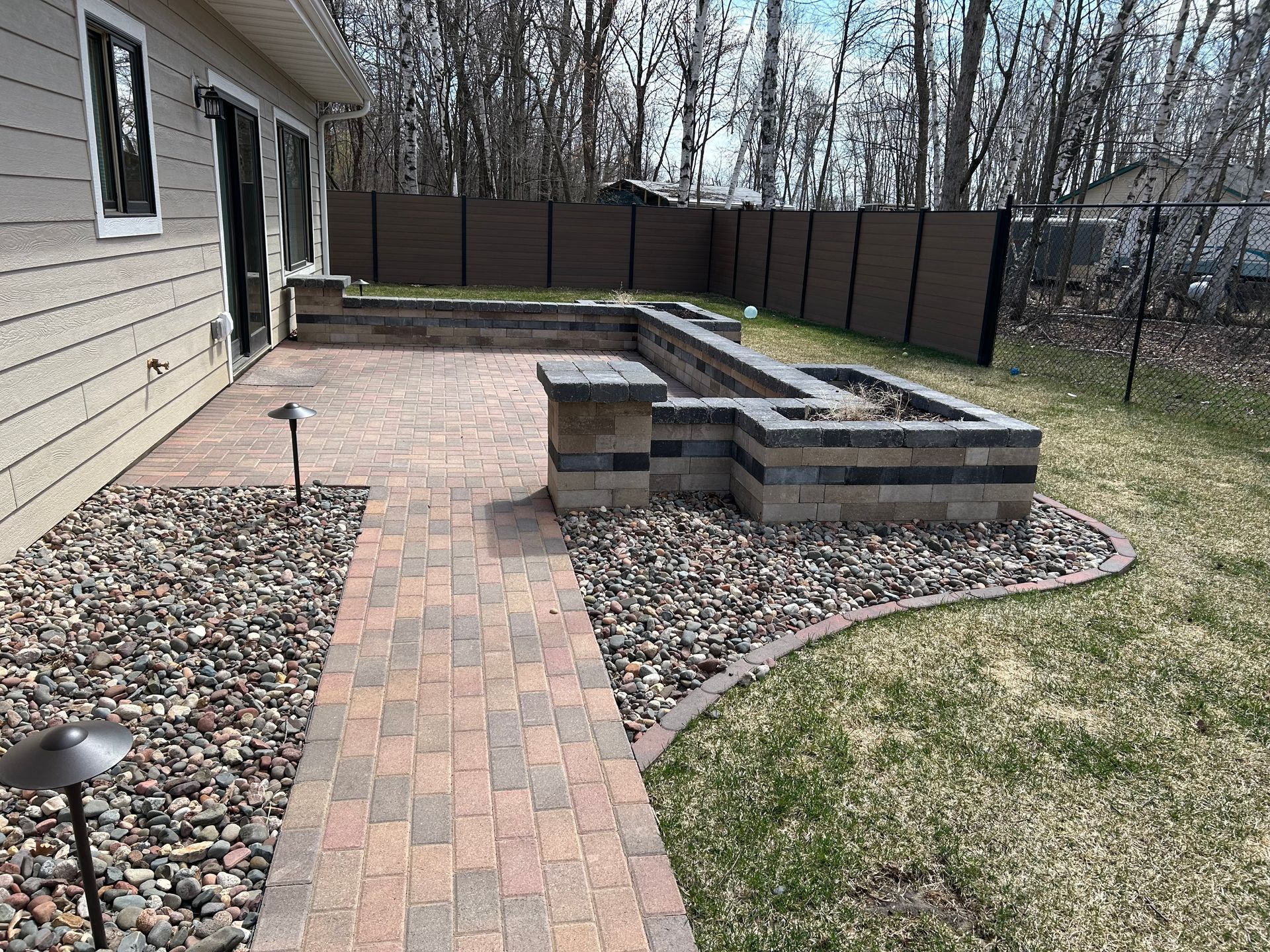 A brick walkway leading to a patio with a fire pit in the backyard of a house.