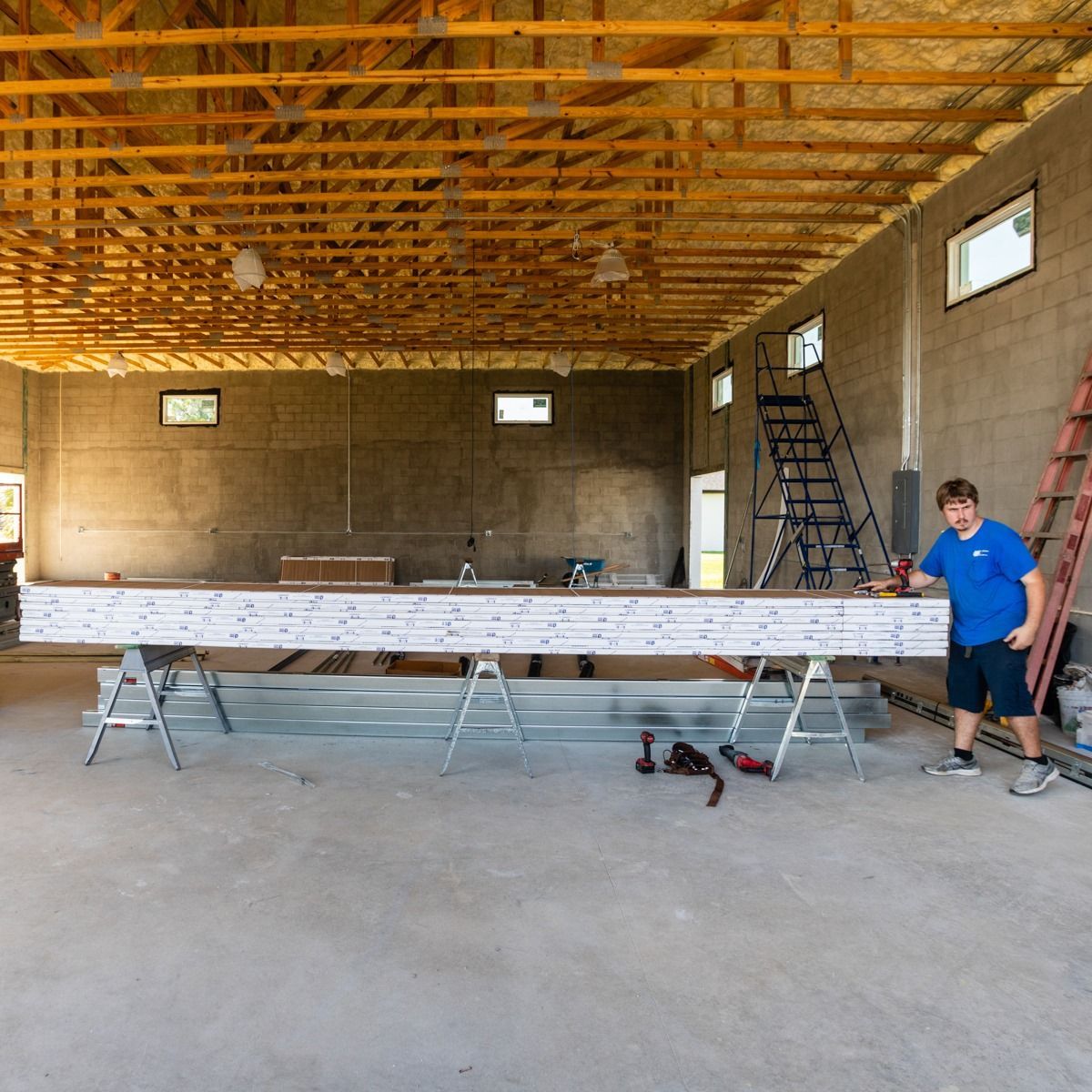 Man measuring long beam in unfinished warehouse. Wooden ceiling, gray walls, tools on floor.
