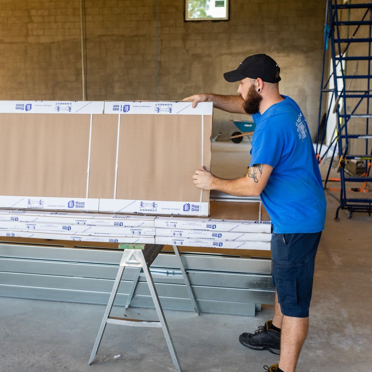 Man in blue shirt and black hat lifting a large packaged window in a construction setting.