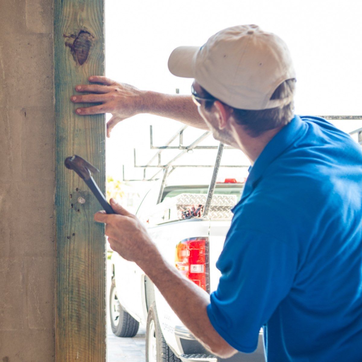 Man in blue shirt and cap hammers wood post near a white truck.