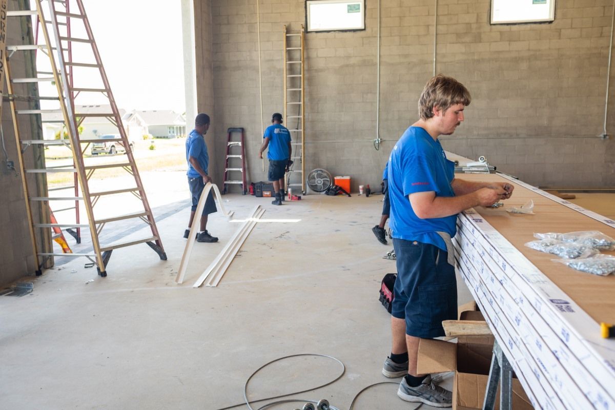 Construction workers installing trim in a large, unfinished building. One worker in focus, others in background.