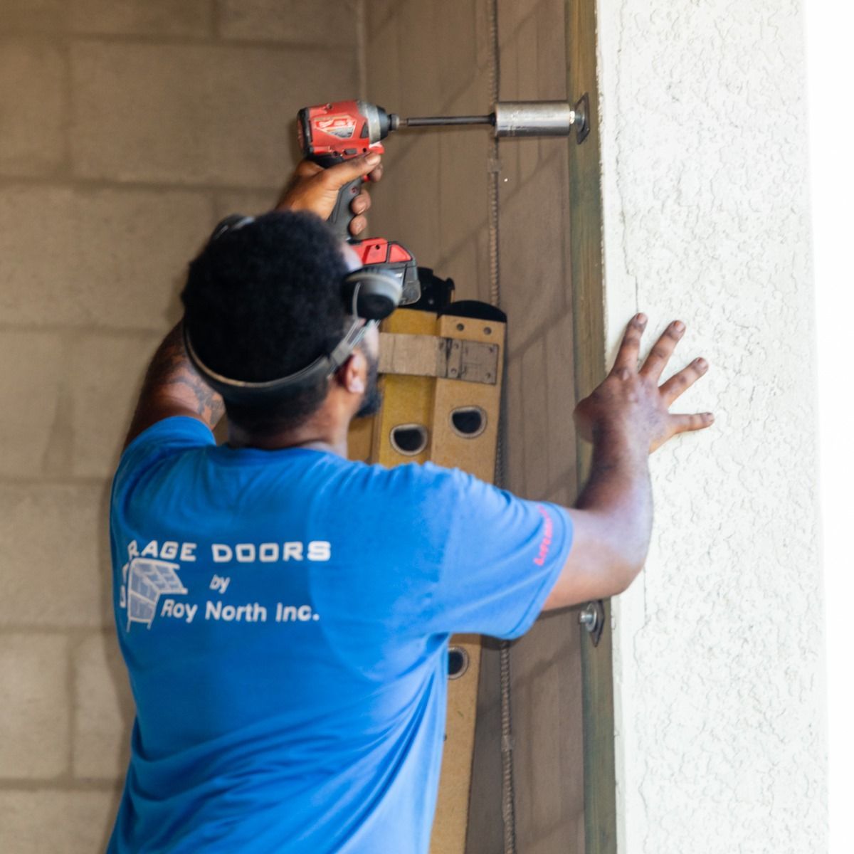 Man in blue shirt using a power drill to install a door, near a ladder and concrete block wall.
