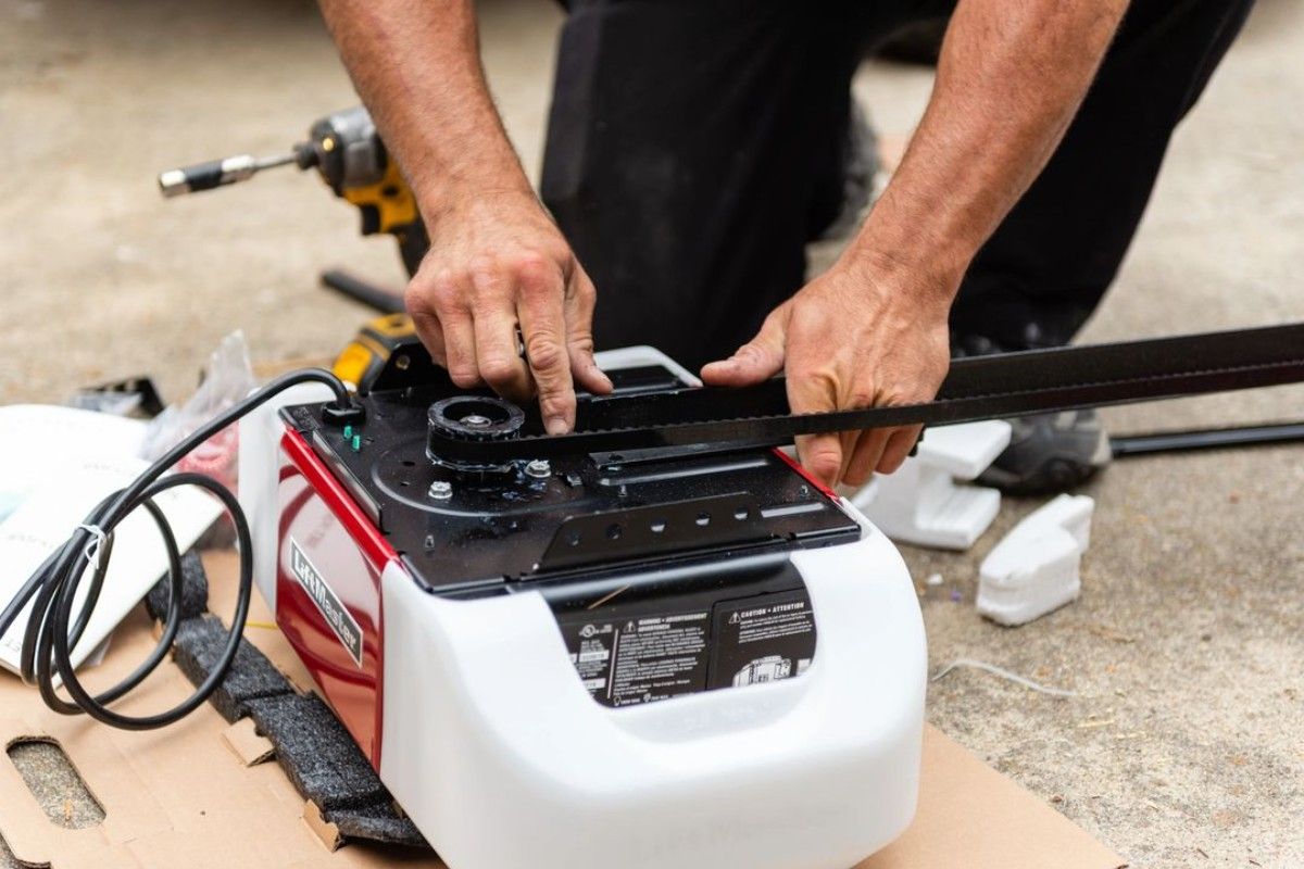 A person installs a garage door opener; red and white unit with a black rail, setting is outdoors.